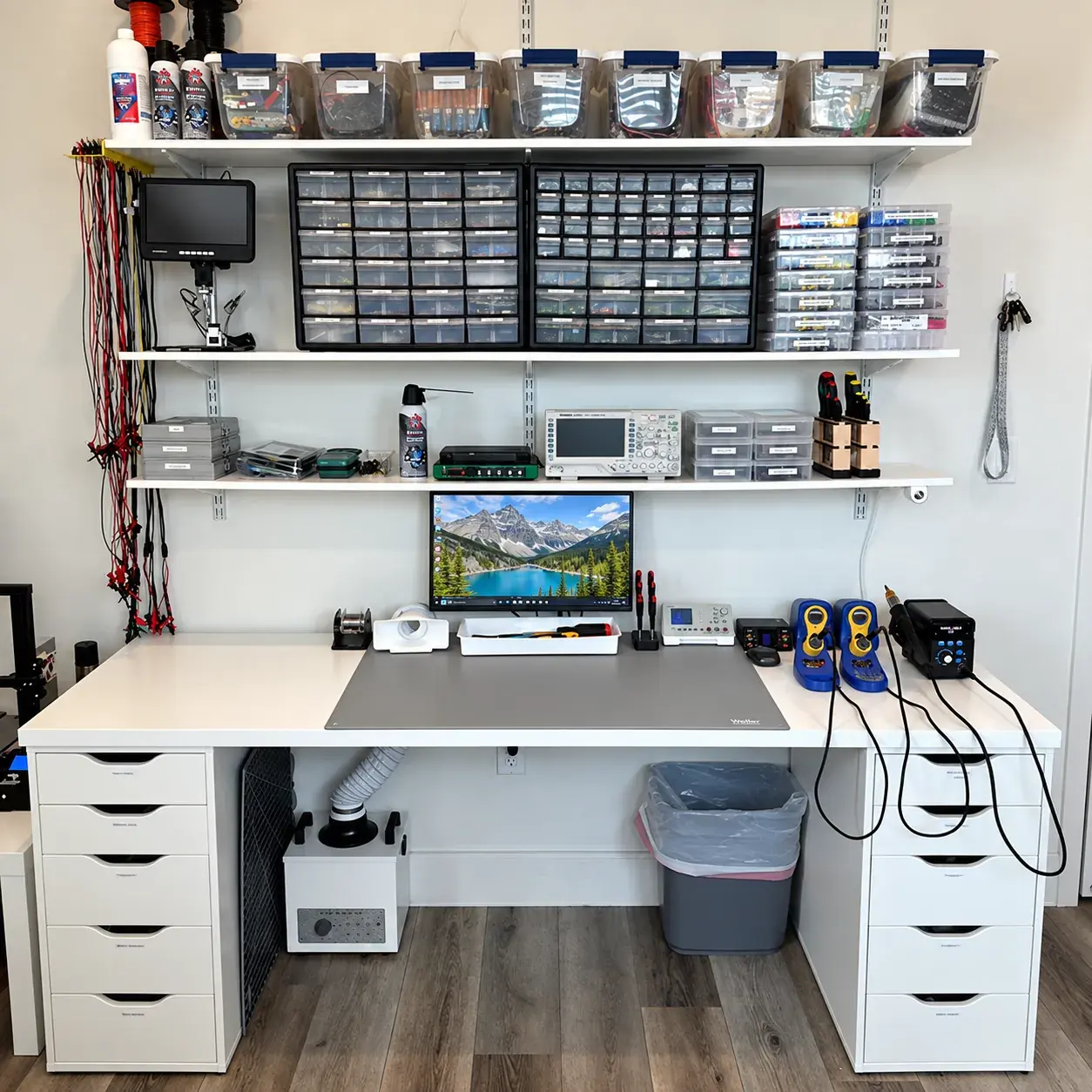 Banco de trabajo de electrónica: mesa blanca con alfombrilla antiestática gris, un monitor, soldadores y una estación de soldadura; encima, estantes con cajas de componentes clasificados, un osciloscopio y un microscopio digital.