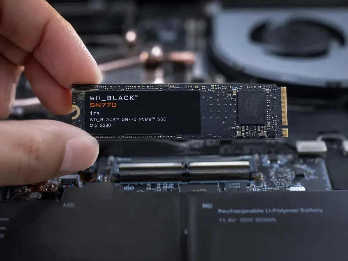 Mano sosteniendo un SSD M.2 NVMe sobre un portátil abierto, con la placa base, el ventilador y la batería a la vista.